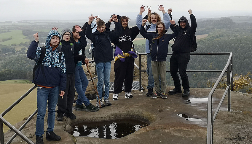 Auf dem Foto ist eine Gruppe abgebildet, die aus sieben Jungen* und zwei Betreuern besteht. Sie befinden sich draußen in der Natur auf einem Aussichtspunkt beim Lilienstein.