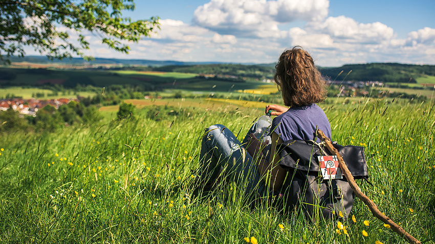 Eine Frau mit Rucksack und Wanderstab sitzt auf einer Wiese.