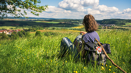 Eine Frau mit Rucksack und Wanderstab sitzt auf einer Wiese.