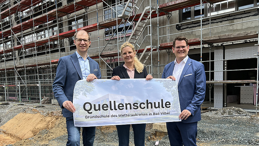 Zwei Männer und eine Frau stehen vor einem Gebäude auf einer Baustelle. Sie halten ein Plakat in den Händen. Darauf steht "Quellenschule".