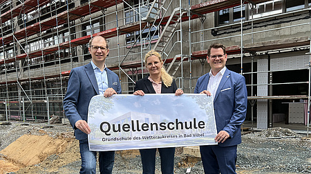 Zwei Männer und eine Frau stehen vor einem Gebäude auf einer Baustelle. Sie halten ein Plakat in den Händen. Darauf steht "Quellenschule".