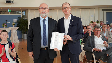 Zwei Männer stehen in einem Saal, im Hintergrund sitzen zahlreiche Menschen. Beide Männer halten eine Urkunde in Händen.