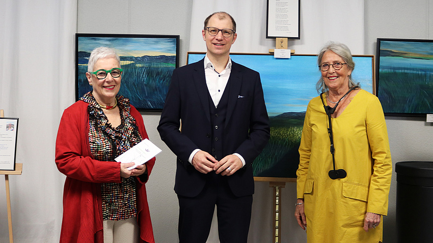 Drei Personen stehen vor einigen Kunstwerken und blicken in die Kamera.