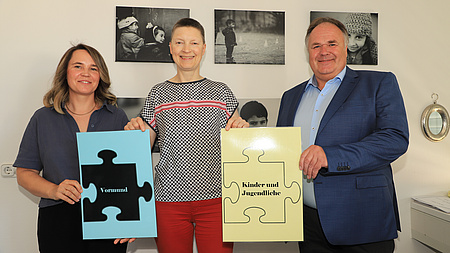 Zwei Frauen und ein Mann stehen in einem Raum vor einer Wand mit Schwarz-Weiß-Fotos von Kindern. Alle drei halten Grafiken von Puzzlesteinen in den Händen.