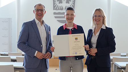 Zwei Frauen und ein Mann stehen in einem Saal. Eine der Frauen hält eine Urkunde in Händen.