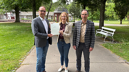 Eine Frau und zwei Männer stehen auf einem Weg in einem Park. Im Hintergrund befindet sich ein Gebäude. Die Frau und einer der Männer halten gemeinsam ein Dokument in Händen.
