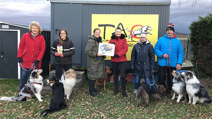 Sechs Menschen mit Hunden stehen vor einem Bauwagen in winterlicher Kulisse.