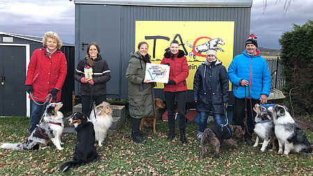 Sechs Menschen mit Hunden stehen vor einem Bauwagen in winterlicher Kulisse.