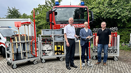 Drei Männer stehen vor einem Feuerwehrauto und zeigen technische Geräte.