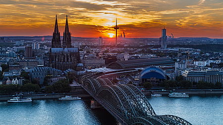Blick auf die Stadt Köln. Im Vordergrund der Rhein, über den eine Brücke führt. In der Mitte der Kölner Dom.