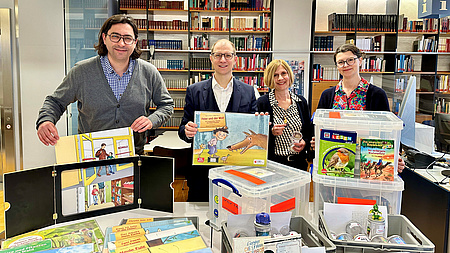Die vier Menschen haben die Bücherkisten vor sich und halten jeweils Medien in die Kamera.
