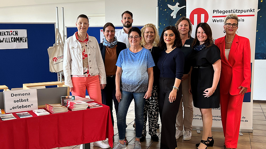 Acht Frauen und ein Mann stehen in einem großen Raum an einem Tisch mit Broschüren vor mehreren Stellwänden.
