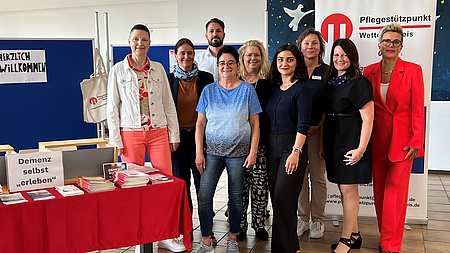 Acht Frauen und ein Mann stehen in einem großen Raum an einem Tisch mit Broschüren vor mehreren Stellwänden.
