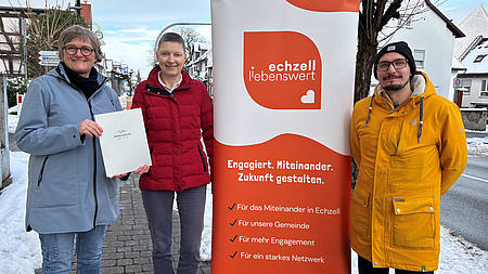 Zwei Frauen und ein Mann stehen im Freien an einer Straße. Zwischen ihnen befindet sich ein Plakat. Im Hintergrund sind Schnee und Wohnhäuser zu sehen. Die beiden Frauen halten gemeinsam ein Dokument in Händen.