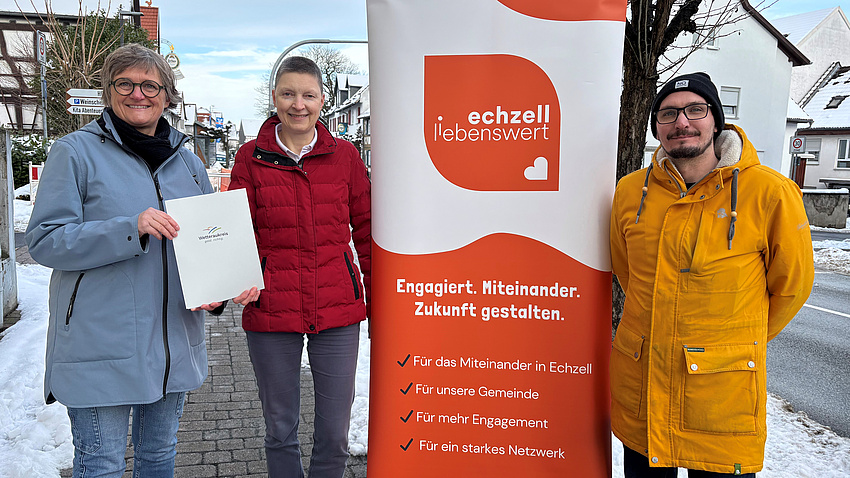 Zwei Frauen und ein Mann stehen im Freien an einer Straße. Zwischen ihnen befindet sich ein Plakat. Im Hintergrund sind Schnee und Wohnhäuser zu sehen. Die beiden Frauen halten gemeinsam ein Dokument in Händen.