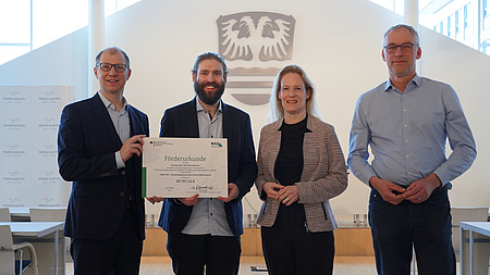 Vier Menschen stehen vor einem Wappen und halten eine Urkunde.