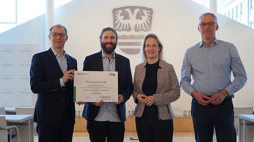 Vier Menschen stehen vor einem Wappen und halten eine Urkunde.