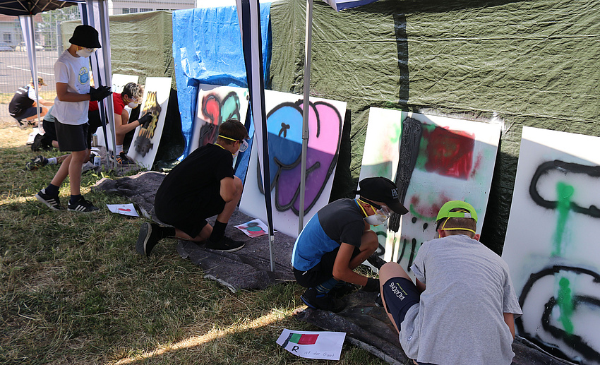 Mehrere Jungen knien oder stehen vor mittelgroßen Holzplatten und besprühen diese mit Farbe. Die Jungen tragen Schutzbrillen und Atemssachutzmasken