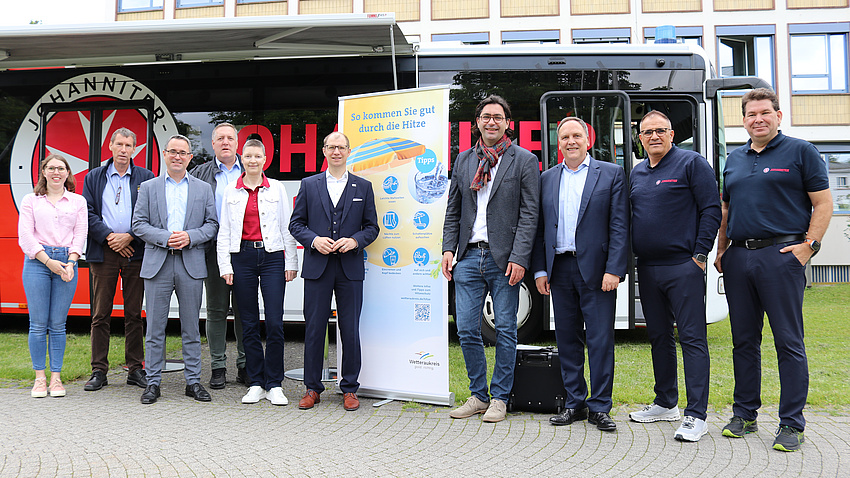 Zehn Männer und Frauen stehen vor einem roten Linienbus. Sie haben sich um ein Roll-Up mit der Aufschrift "So kommen Sie gut durch die Hitze" gruppiert. Sie lächeln in die Kamera.