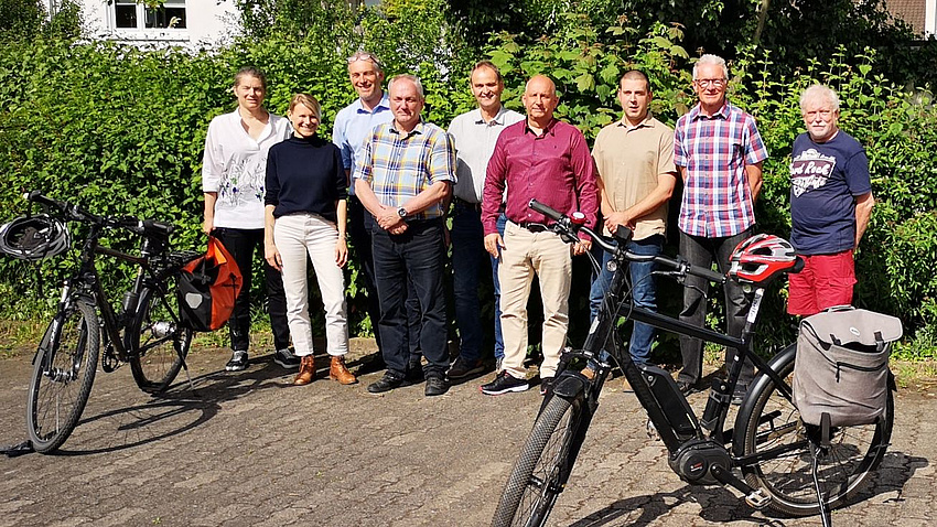 Eine Gruppe von Leuten steht vor einer grünen Hecke. Zwei Fahrräder stehen neben ihnen.