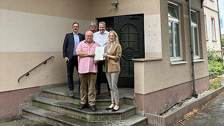 Ein Frau und vier Männer stehen auf einer Treppe vor einem Hauseingang. Die Frau übergibt einem der Männer ein Dokument.