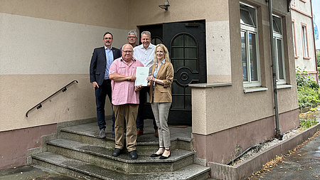 Ein Frau und vier Männer stehen auf einer Treppe vor einem Hauseingang. Die Frau übergibt einem der Männer ein Dokument.