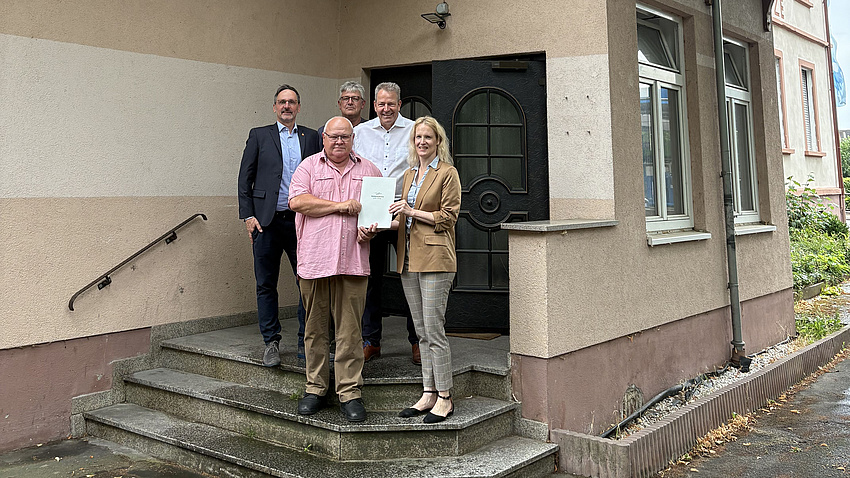 Ein Frau und vier Männer stehen auf einer Treppe vor einem Hauseingang. Die Frau übergibt einem der Männer ein Dokument.