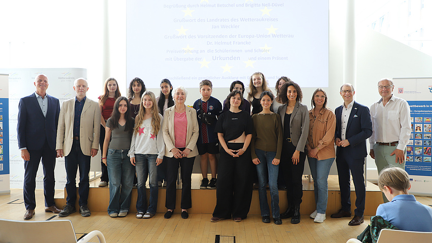 Einige ältere Schülerinnen und Schüler stehen gemeinsam mit einigen Erwachsenen in einem großen Saal. Im Hintergrund sieht man ein Plakat und eine PowerPoint-Präsentation auf einer Leinwand.