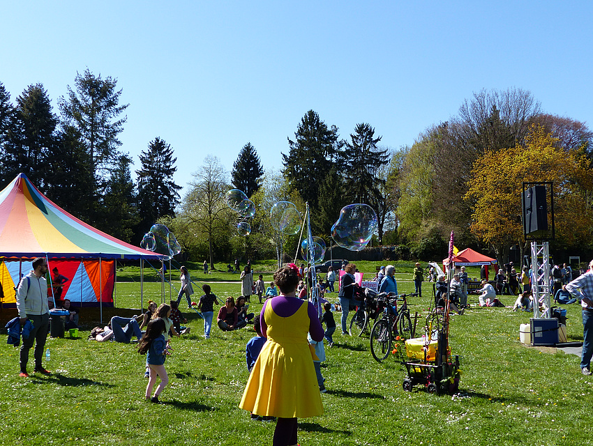 Auf Wiese stehen Menschen und Kinder spielen, dahinter Bäume