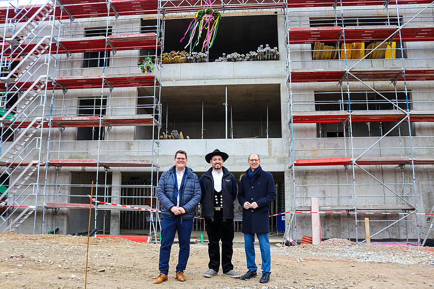 Drei Männer stehen vor einem Rohbau und lächeln in die Kamera. Einer von ihnen trägt eine traditionelle Handwerkerkluft.