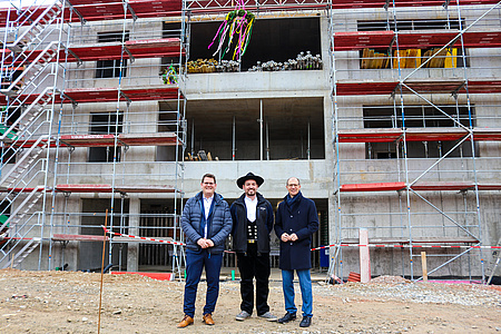 Drei Männer stehen vor einem Rohbau und lächeln in die Kamera. Einer von ihnen trägt eine traditionelle Handwerkerkluft.