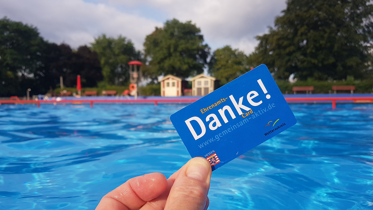 eine Hand hält die blaue Ehrenamtscard mit der Aufschrift Danke, dahinter das Wasser eines Freibades