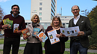Zwei Frauen und zwei Männer stehen auf einem Platz im Freien, im Hintergrund ist ein Hochhaus zu sehen. Alle vier halten Bücher und Flyer in Händen.