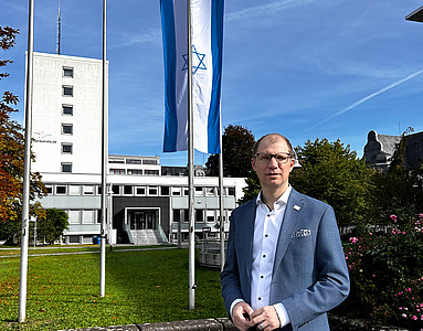 Ein Mann steht auf einem freien Platz. Im Hintergrund weht an einem Fahnenmast die israelische Nationalflagge.
