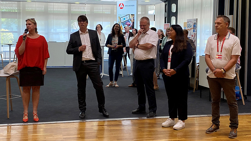 Zwei Frauen und drei Männer stehen in leichtem Abstand nebeneinander. Eine Frau spricht in ein Mikrofon.