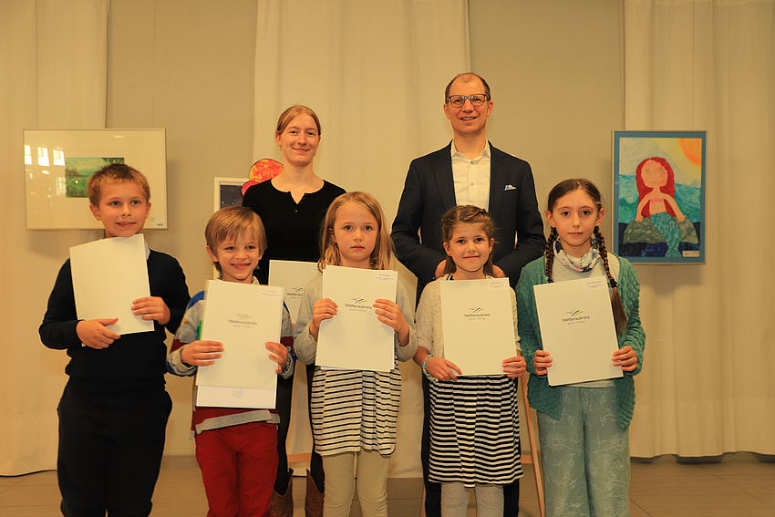 Ein Erwachsener, eine Jugendliche und fünf Kinder, zwei Jungen und drei Mädchen, stehen in einem Raum vor mehreren Bildern. Die Kinder und die Jugendliche halten Urkunden in den Händen.