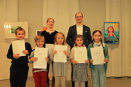 Ein Erwachsener, eine Jugendliche und fünf Kinder, zwei Jungen und drei Mädchen, stehen in einem Raum vor mehreren Bildern. Die Kinder und die Jugendliche halten Urkunden in den Händen.