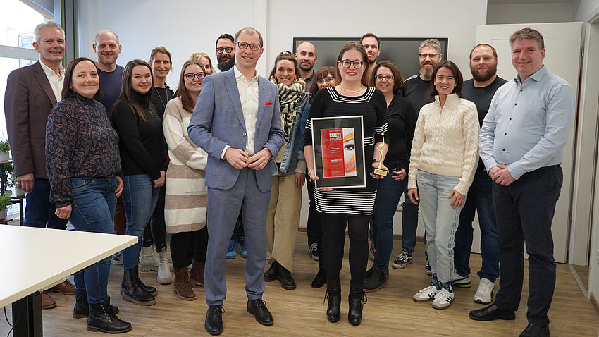 Das Foto zeigt das Team des Fachdienstes Schul-IT und Ausstattung. Sie stehen gemeinsam in einem Büro, die Vorgesetzte Steht mit dem Landrat vorne und zeigt die Urkunde in die Kamera.