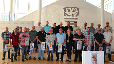Zahlreiche Männer und Frauen stehen mit Wimpeln in der Hand vor einer Fensterfront mit einem Wappen.