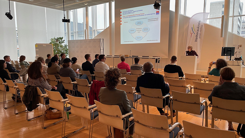 Mehrere Personen sitzen in einem Saal, während eine Frau an einem Rednerpult einen Vortrag hält.