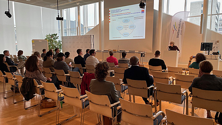 Mehrere Personen sitzen in einem Saal, während eine Frau an einem Rednerpult einen Vortrag hält.