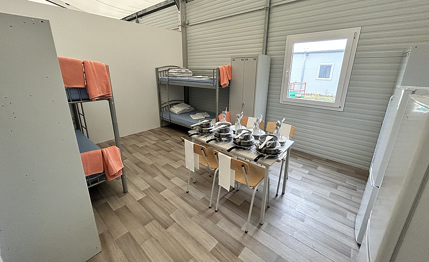 Ein mit einer Trennwand abgetrennter Raum in einer Halle. Darin zwei Stockbetten, zwei Kühlschränke, ein Tisch mit vier Stühlnen. Auf dem Tisch ist Kochgeschirr