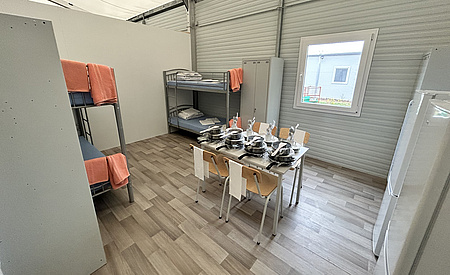 Ein mit einer Trennwand abgetrennter Raum in einer Halle. Darin zwei Stockbetten, zwei Kühlschränke, ein Tisch mit vier Stühlnen. Auf dem Tisch ist Kochgeschirr