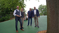 Vier Personen stehen auf einer neu gestalteten Spielfläche an einem Hang. Der versickerungsfähige Belag ist grün,  im Hintergrund ist eine Rutsche zu sehen.