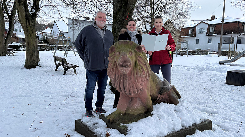 Zwei Frauen und ein Mann stehen in einem verschneiten Parkgelände hinter der Sandstein-Skulptur eines Löwen. Die beiden Frauen halten gemeinsam ein Dokument in Händen.
