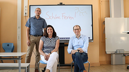Drei Erwachsene sitzen auf Stühlen für Grundschüler vor einer digitalen Tafel.