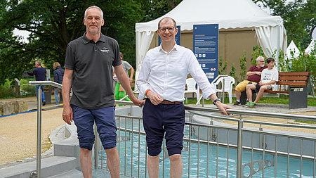 Zwei Männer stehen im Freien mit nackten Füßen im Wasser eines Kneipp-Beckens und halten sich an einem Geländer fest.
