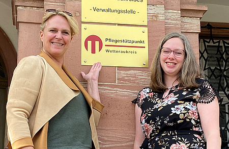 Zwei Frauen stehen vor einem Gebäude. Eine der beiden deutet auf ein Schild, auf dem steht: Pflegestützpunkt Wetteraukreis
