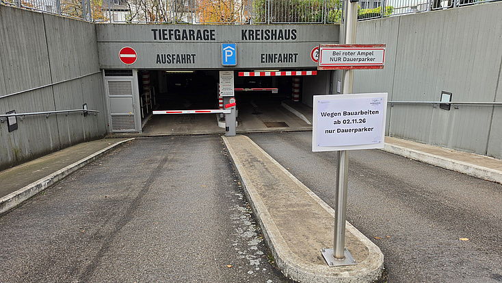 Zweispurige Zufahrt zu einer Tiefgarage mit Schranken, an der ein Hinweisschild angebracht ist.
