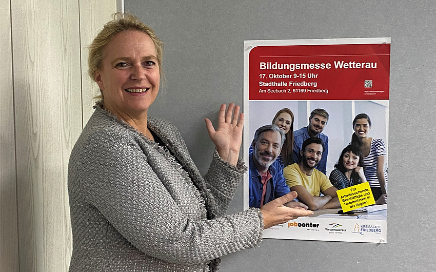 Eine Frau weist auf ein Plakat an einer Stellwand. Das Plakat wirbt für die Bildungsmesse.